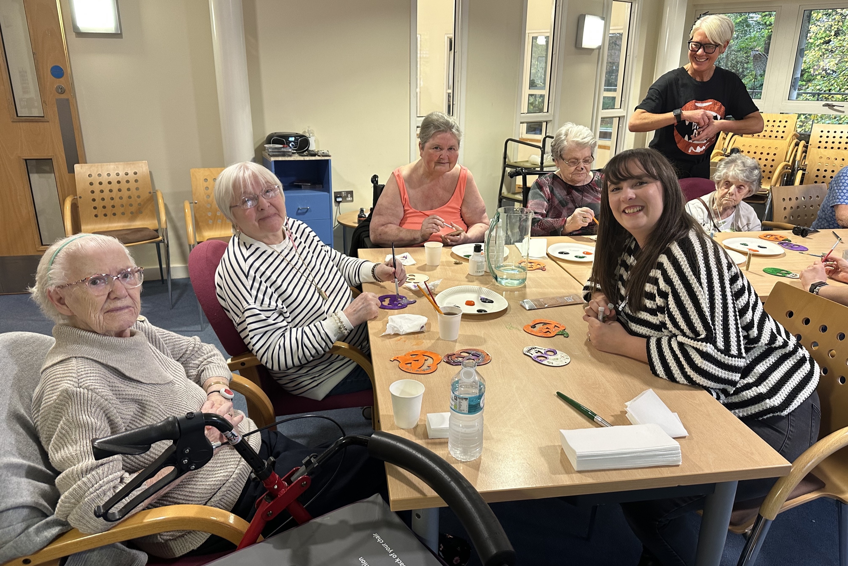 A group of older adults and a younger woman sit around a table, painting Halloween decorations like pumpkins and skulls, smiling at the camera.