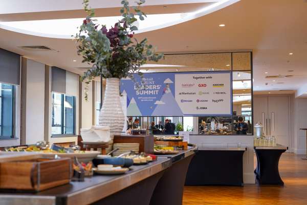 Event buffet with snacks and drinks. In the background, a screen shows 'Retail Trust Leaders' Summit' and sponsor logos. Attendees mingle.