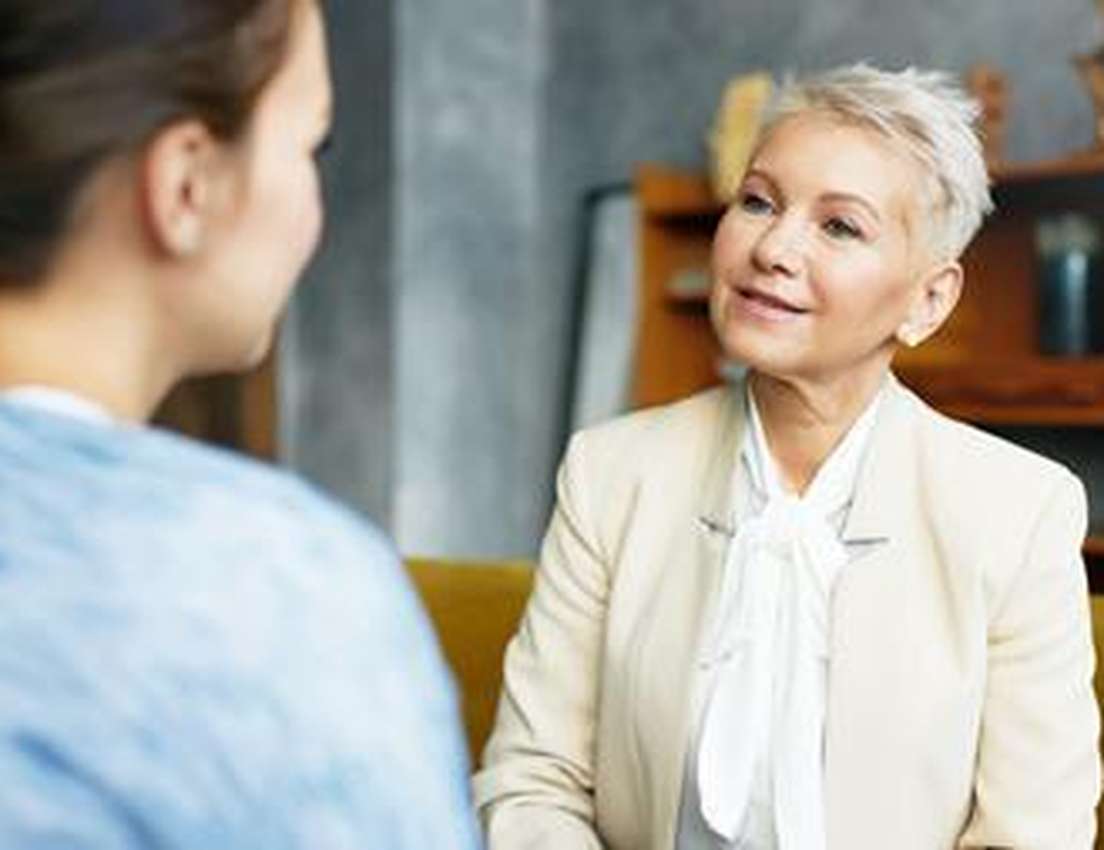 An older woman with short white hair in a cream blazer smiles, listening to a younger woman with her back to the viewer.