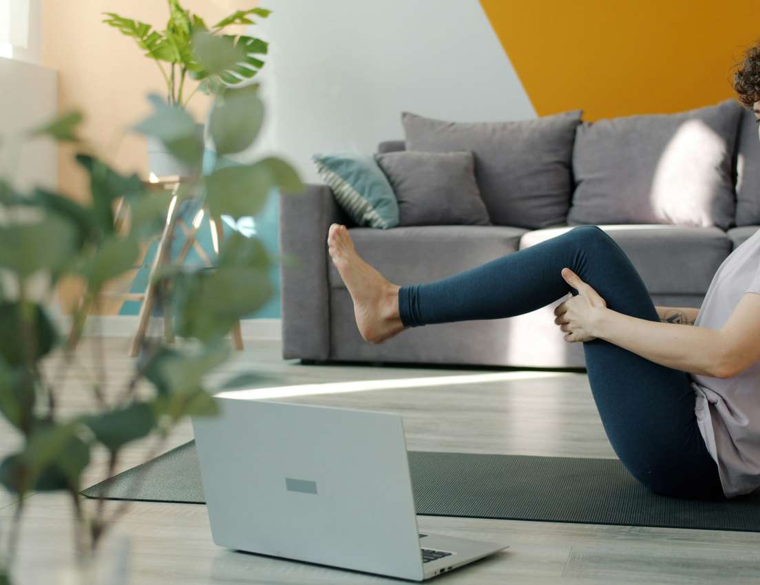 A woman practices boat pose on a yoga mat, following an online class on her laptop in a modern living room.