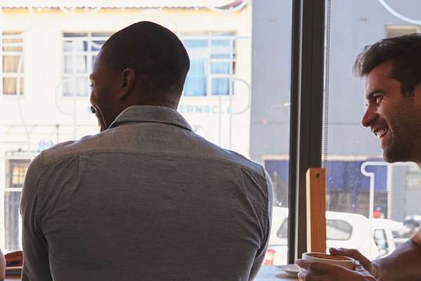 Three diverse men smiling and talking in a bright cafe, one seen from behind.