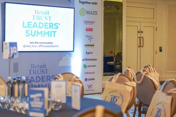 Empty conference room for "Retail Trust Leaders' Summit" with screen, sponsor banner, tables, and chairs topped with branded 