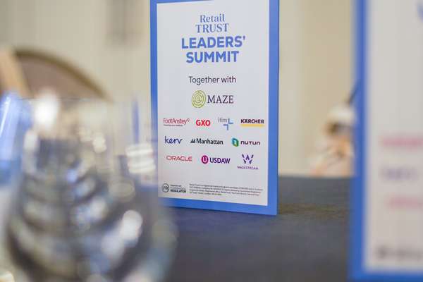 White sign for "Retail TRUST LEADERS' SUMMIT" with sponsor logos, on a table. A blurry glass is visible in the foreground.