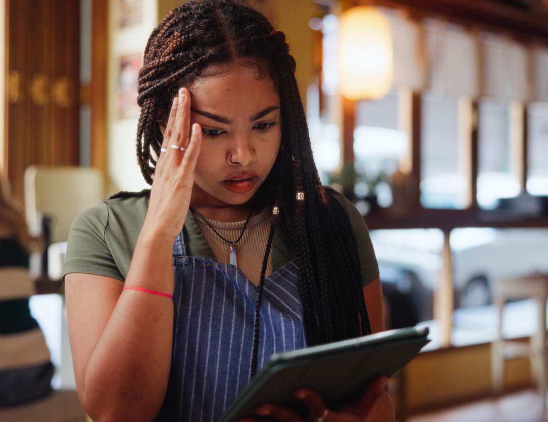 A young woman with braided hair, wearing a cafe apron, looks worried while holding a tablet, with her hand on her forehead.