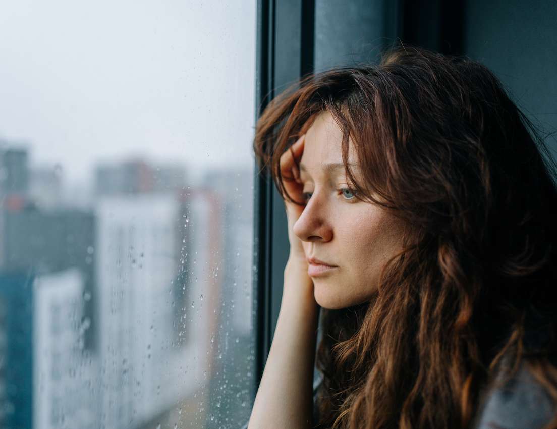 Close-up of a thoughtful woman with long brown hair, looking out a rain-covered window at a blurred city landscape.