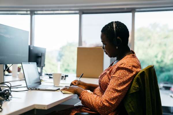 A Black woman wearing headphones writes in a notebook at an office desk with a laptop and monitors, large windows behind her.