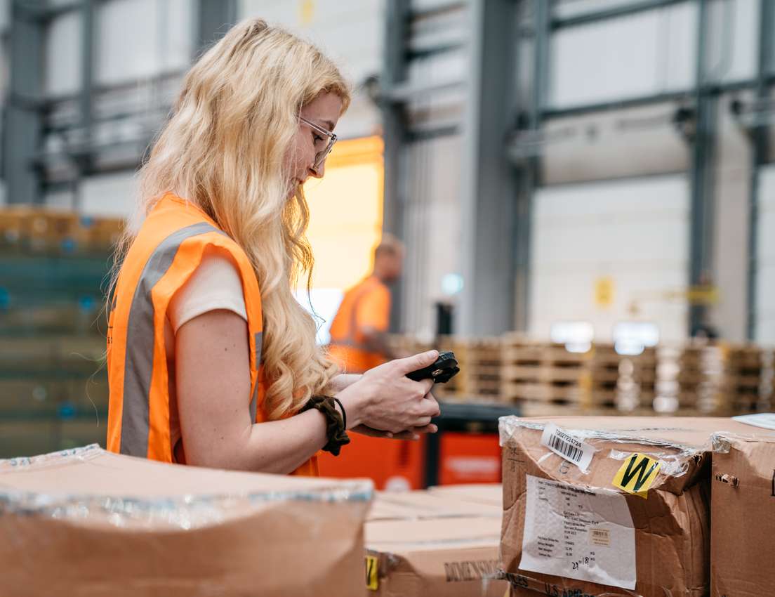A blonde woman in an orange safety vest scans a package with a handheld device in a busy warehouse. Cardboard boxes are stack