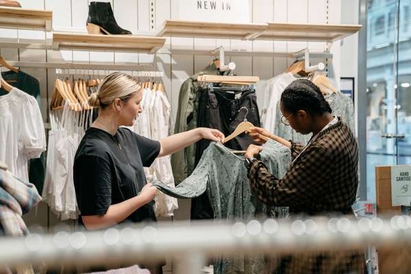 A blonde sales assistant presents a green floral dress to a Black woman in glasses, browsing clothes in a bright retail store.