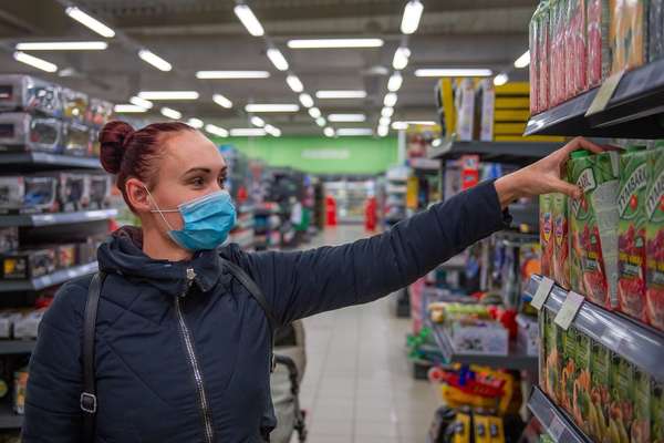 Woman in a blue surgical mask and dark quilted jacket reaching for a carton of juice on a shelf in a brightly lit supermarket.
