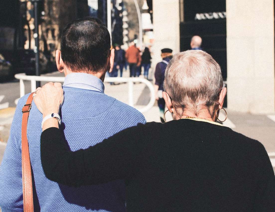 Man and woman walking away, her arm around his shoulder. He has dark hair, blue sweater; she has gray hair, black top, gold h