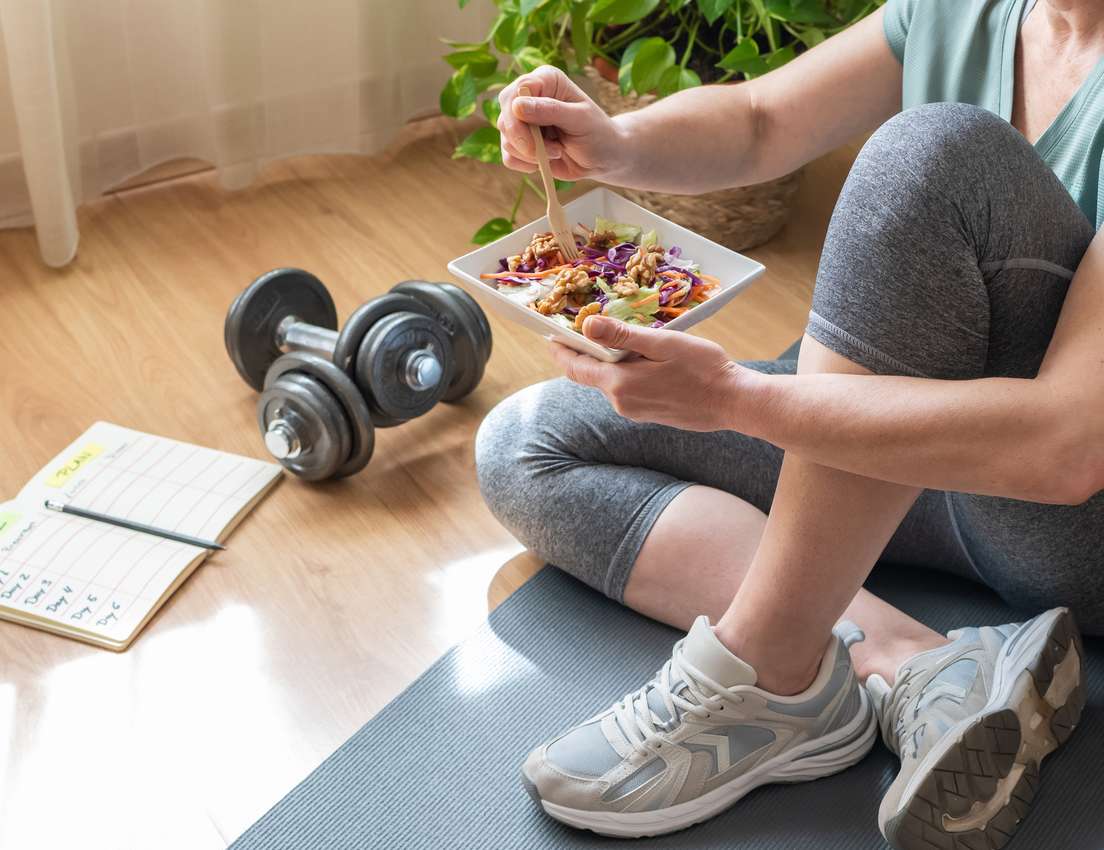 A person in workout clothes sits on a mat, eating a colorful salad with walnuts. Dumbbells and a meal plan notebook are on th