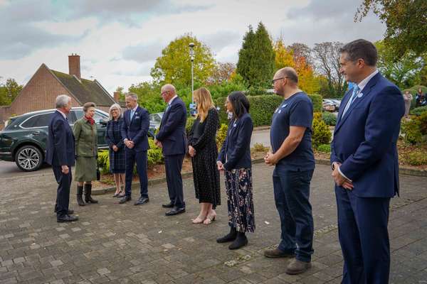Princess Anne, in green tweed, greets a line of men and women outdoors. An SUV, brick building, and autumn trees are in the b