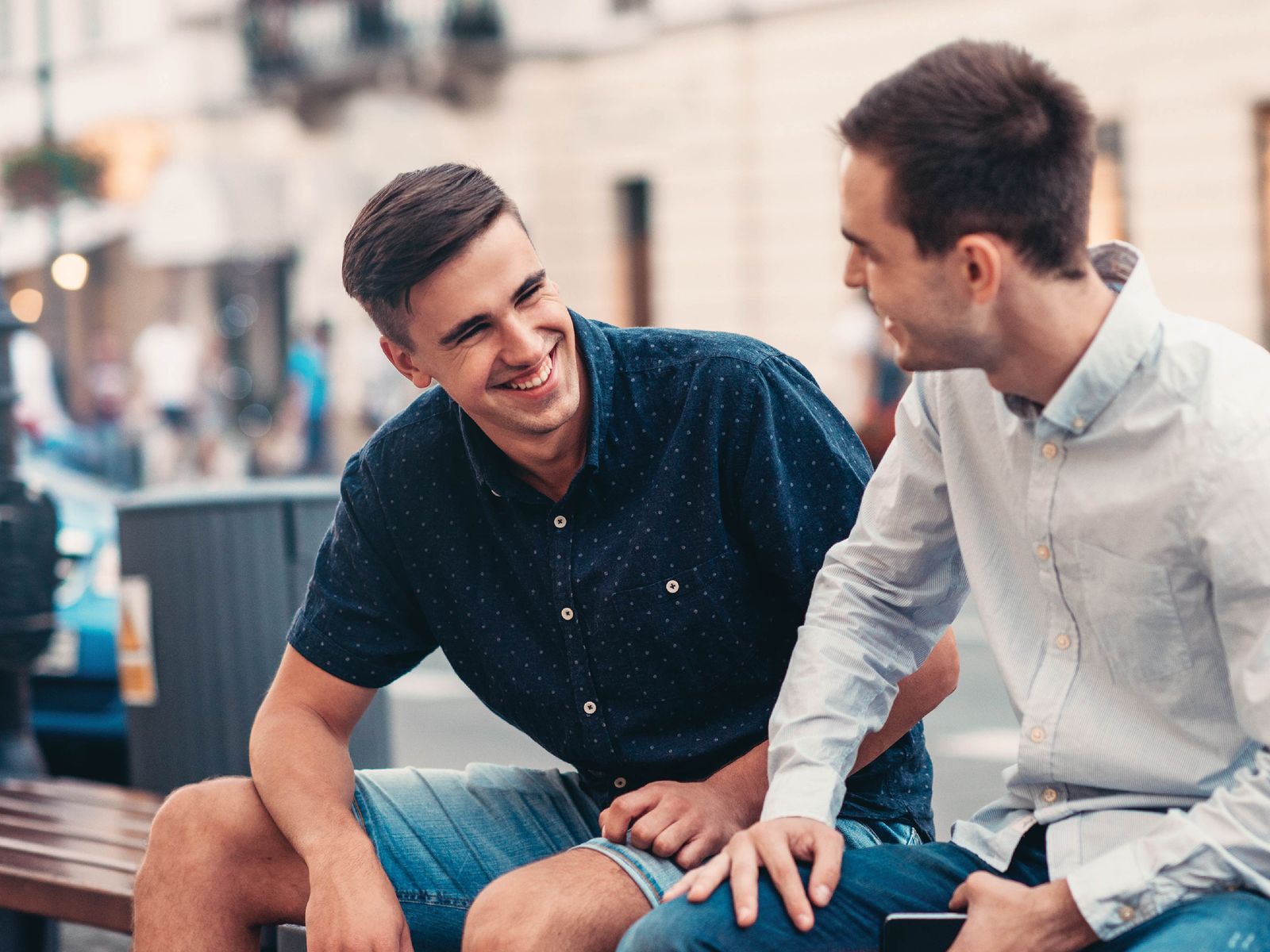 Two men sitting on a bench, smiling and engaging in conversation on a city street.