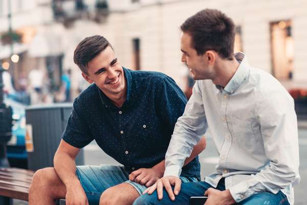 Two men sitting on a bench, smiling and engaging in conversation on a city street.