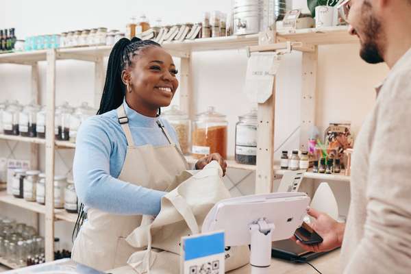 Smiling Black woman in an apron packs a reusable bag for a customer paying with a phone at a bulk store.