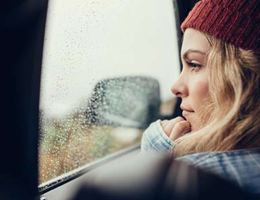 Woman in a red beanie looks thoughtfully out a rainy car window, resting her chin on her hand.