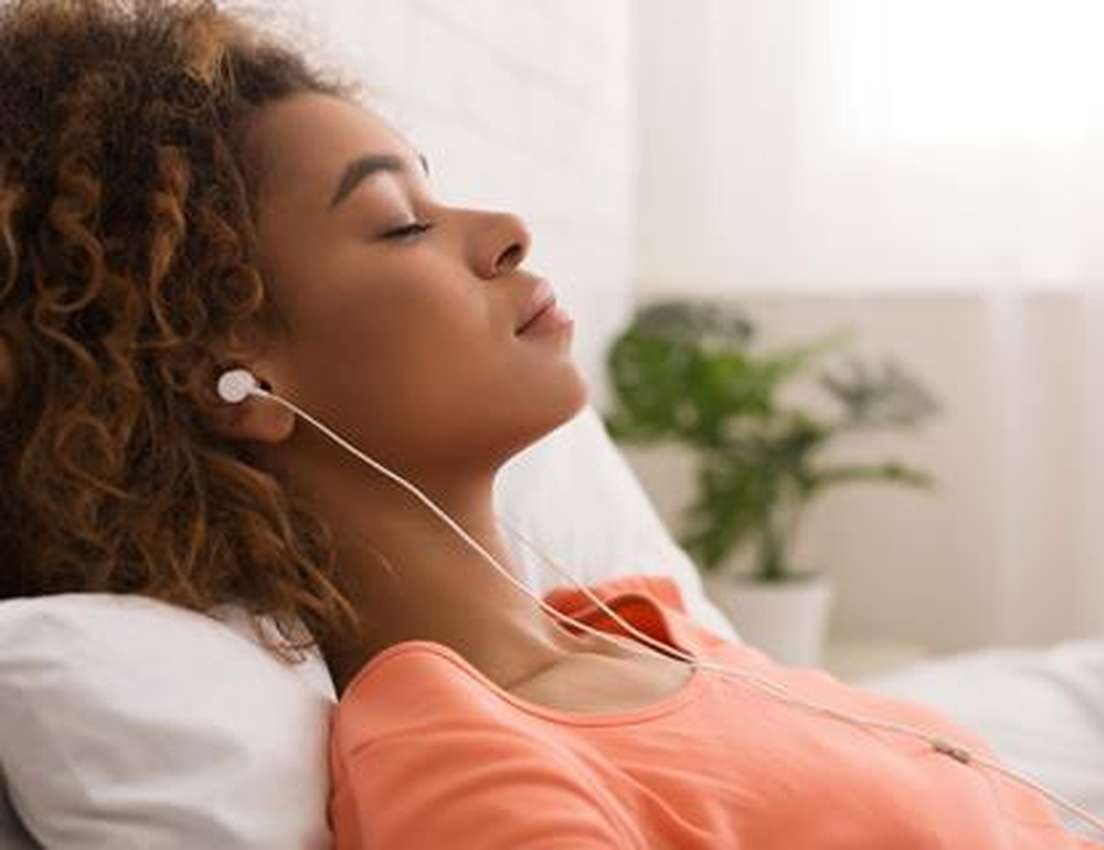A person with curly hair relaxes on a couch, eyes closed, listening to music with earphones. There's a potted plant in the ba