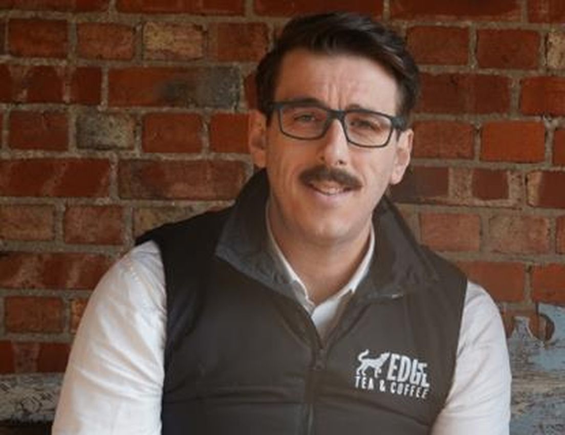 Man with a mustache and glasses, smiling in a black Edge Tea & Coffee vest against a brick wall.