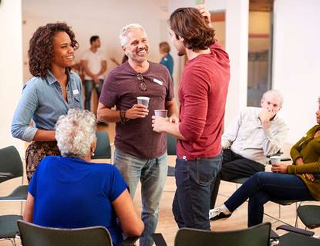 People conversing and socializing in a casual setting, with chairs arranged in a circle and some holding cups, suggesting a g