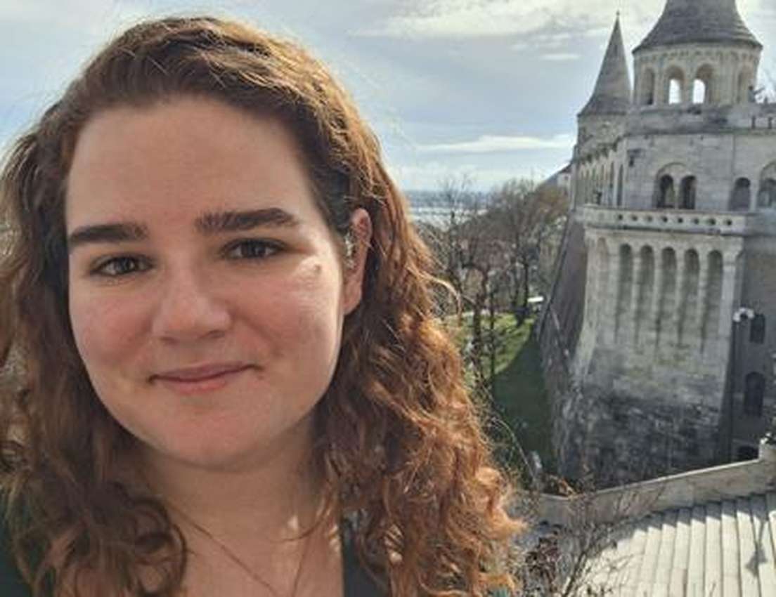 Smiling woman with curly hair in front of Budapest's Fisherman's Bastion with a city vista.