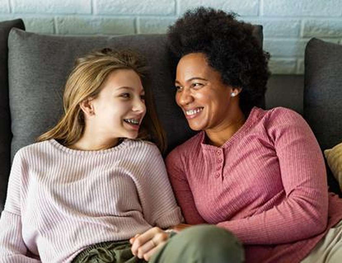 A happy Black woman with an afro and a blonde teen with braces smile at each other while relaxing on a couch.