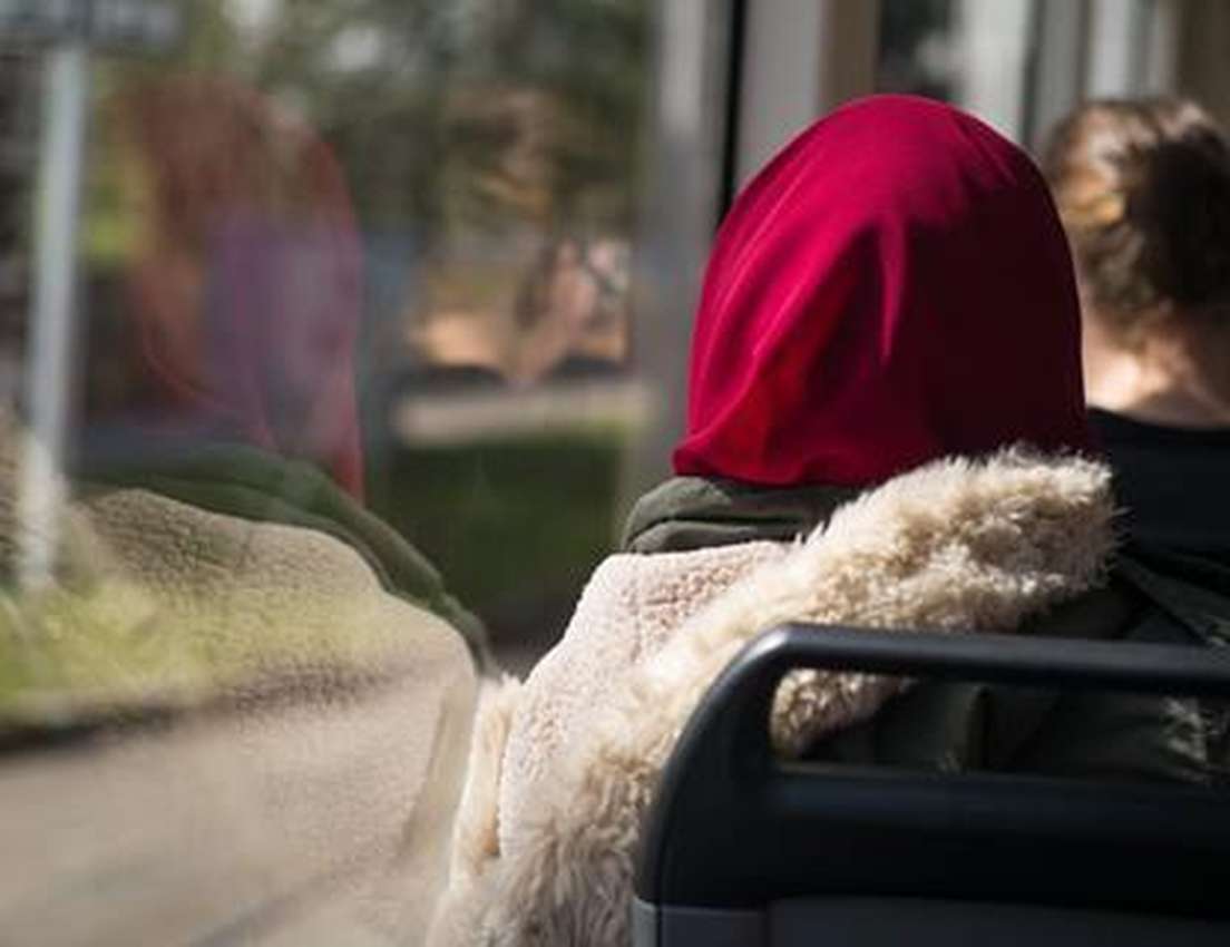 Back of person in red head covering & fluffy jacket, seated by window showing their reflection & blurred scenery outside.