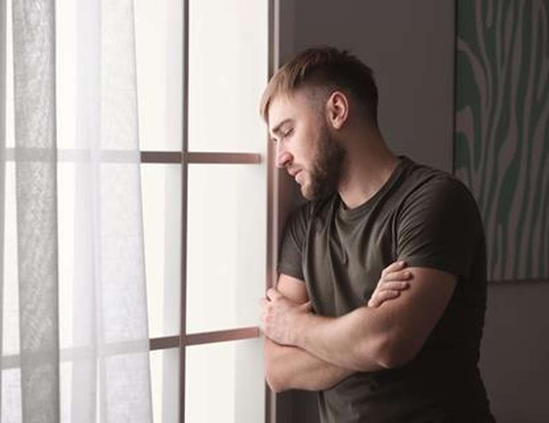Pensive bearded man with arms crossed leans against a window, looking down.