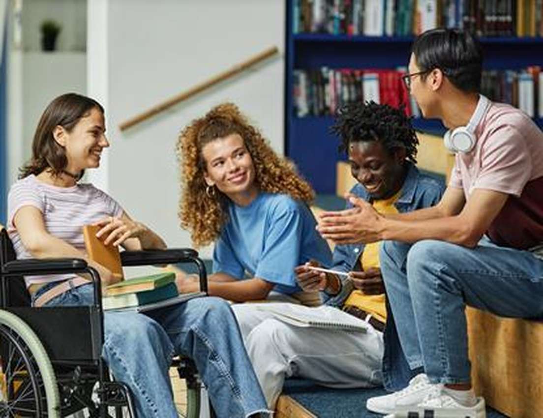Four diverse students, one in a wheelchair, smiling and engaged in conversation in a library.