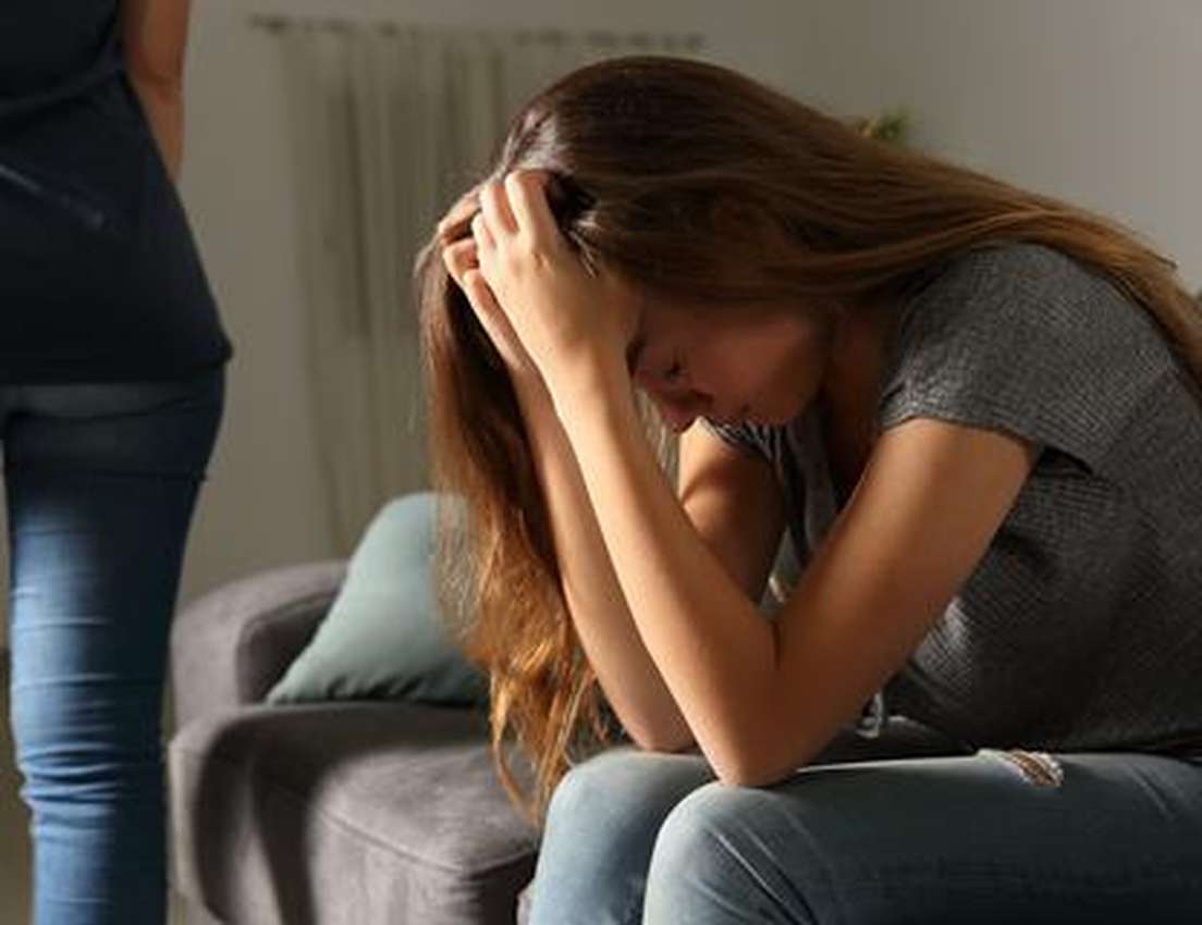 A distraught woman sits, head in hands, while another person stands with their back to her.