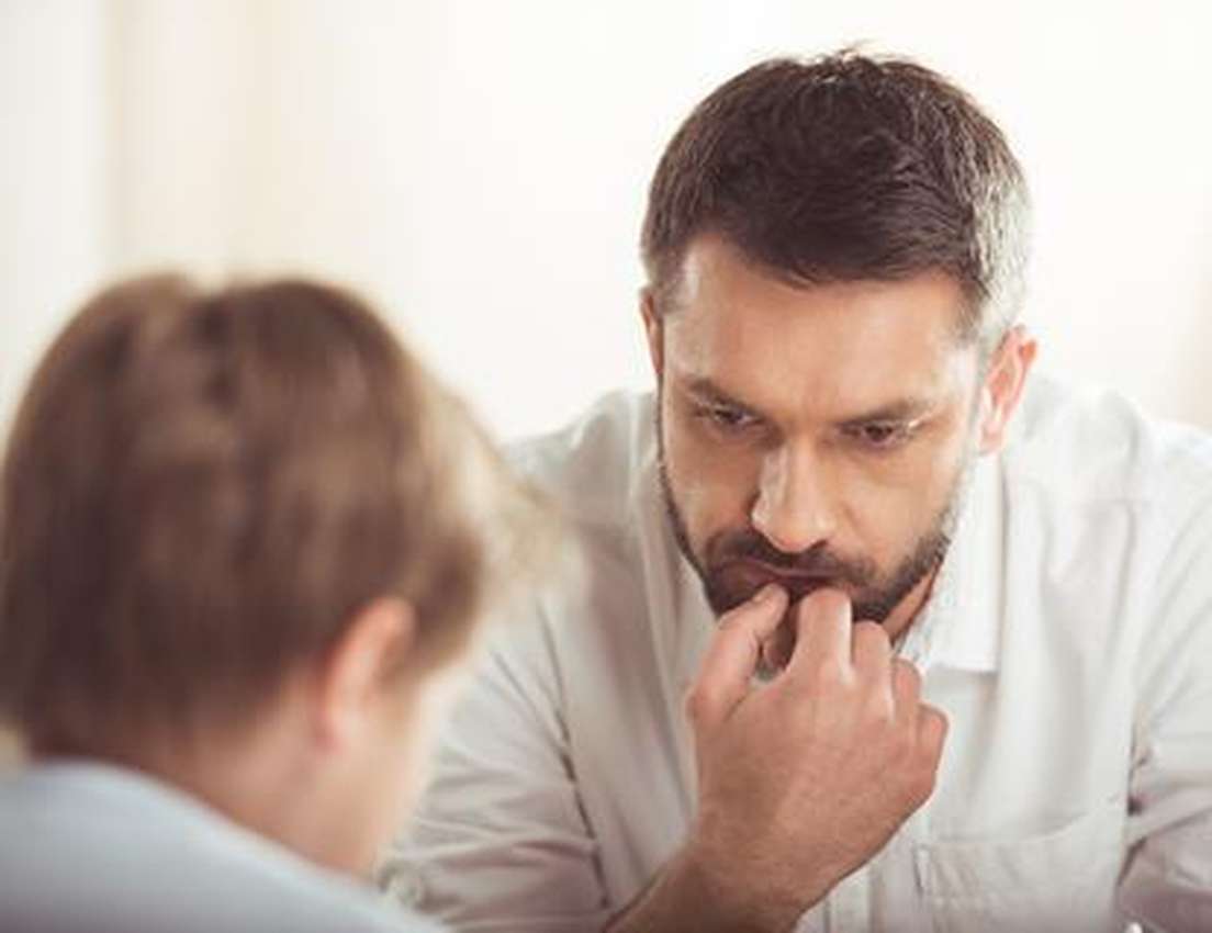 An anxious man in a white shirt bites his nails while looking down intently at a blurred child in the foreground.