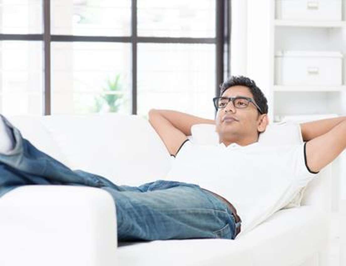 Man in glasses lounging on a white sofa, hands behind head, wearing a white shirt and jeans, with a bright window in the back