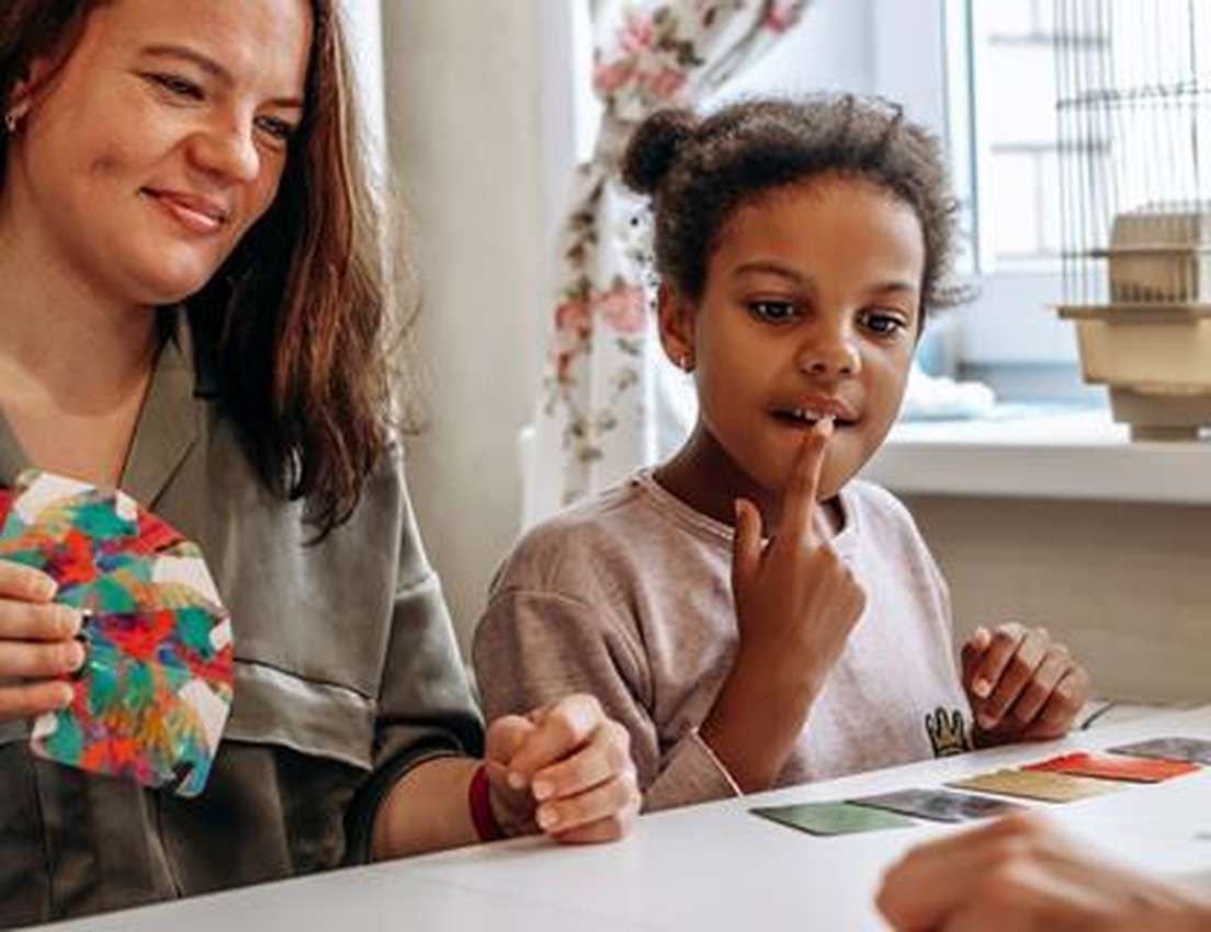 A smiling woman and a thoughtful young girl playing cards at a table, with a birdcage in the background.