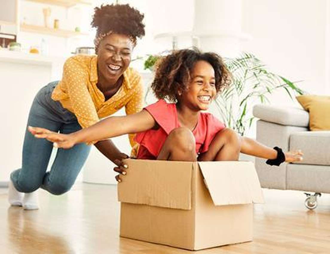 Joyful Black woman pushes a laughing Black child in a cardboard box across a bright room.