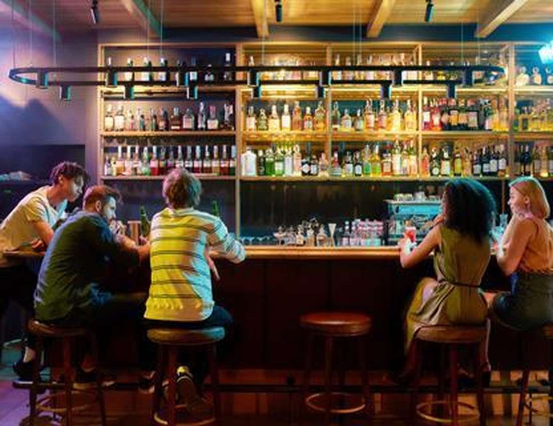 Four people enjoying drinks at a lively bar, two men standing and two women sitting, with a large illuminated liquor shelf be