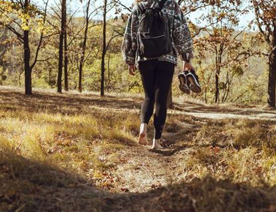 Person walking barefoot on a grassy forest path, holding shoes, wearing leggings and a patterned sweater with a backpack.