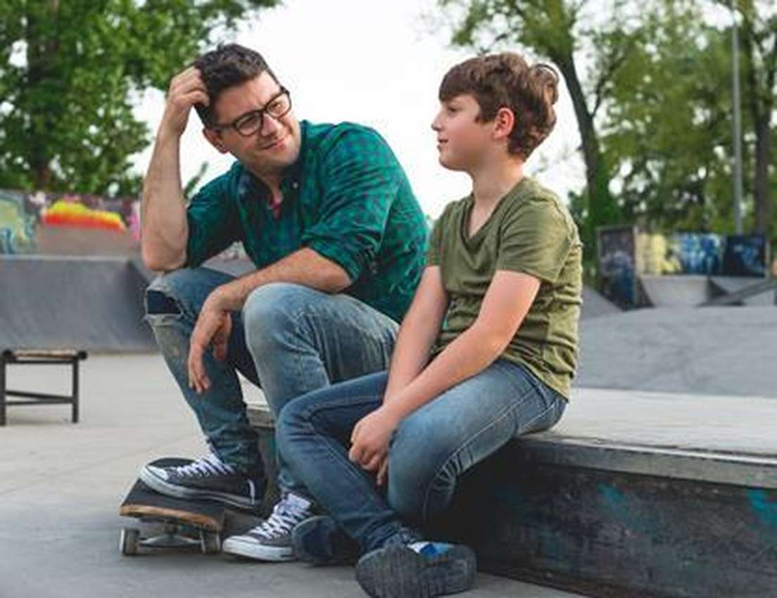 Smiling man in glasses and boy sit on a skatepark ledge. The man looks at the boy, with a skateboard near his feet.