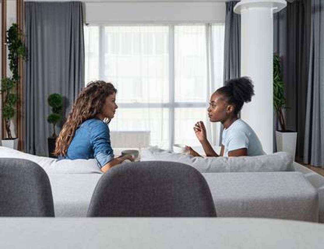 Two women sitting on a sofa, engaged in conversation in a modern living room with large windows and gray curtains.