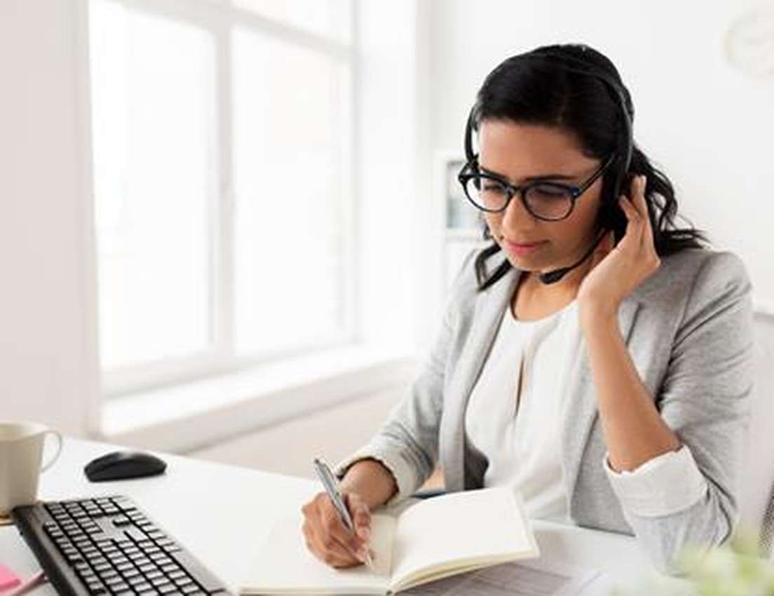 Woman in glasses wearing a headset, writing in a notebook at a desk with a computer, keyboard, and coffee cup in a bright off