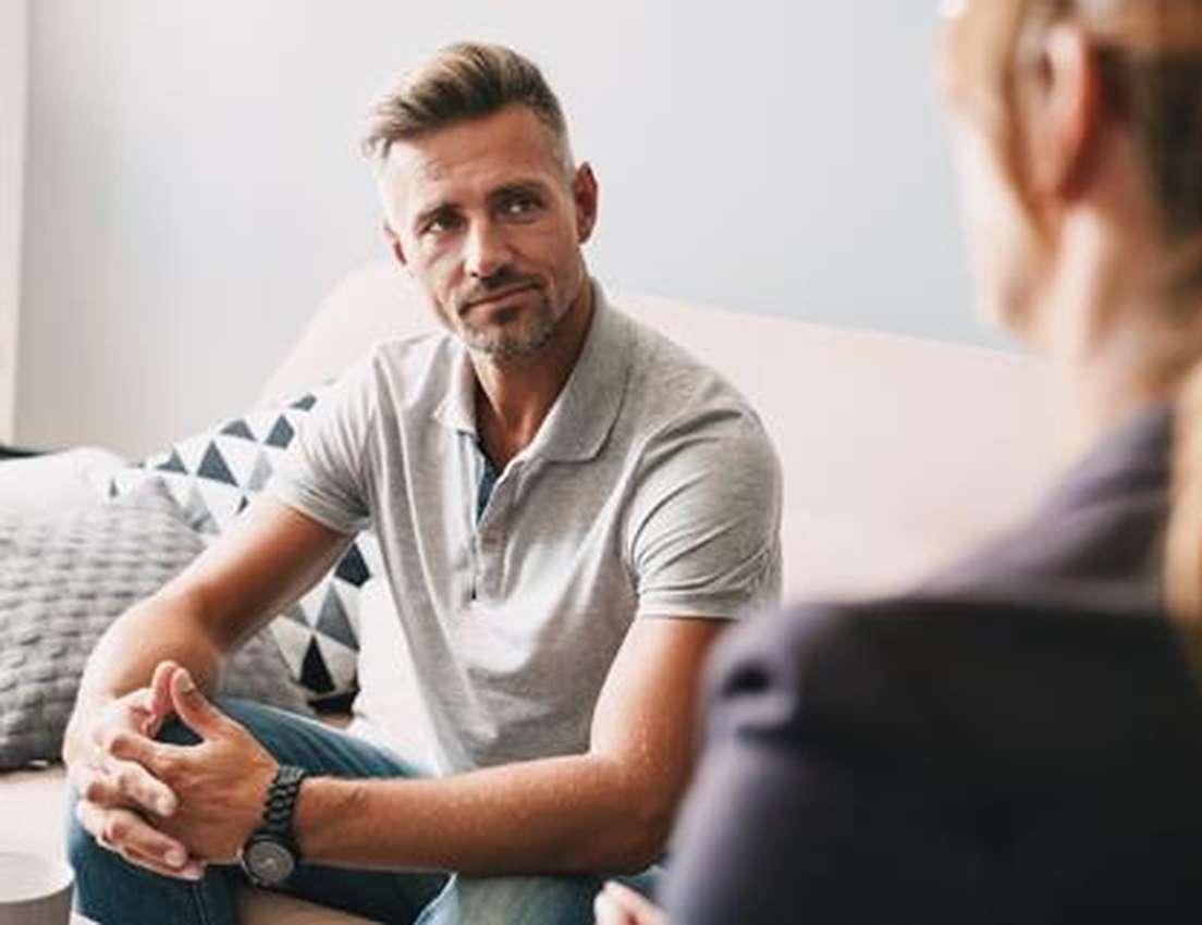 Man in a grey polo shirt sitting on a sofa, attentively listening to a woman during a conversation in a cozy room.
