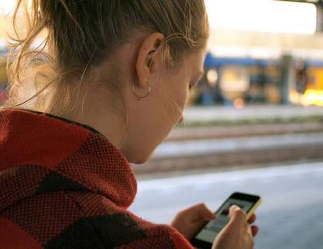 Woman in red plaid hoodie looking at her smartphone at a train station platform.