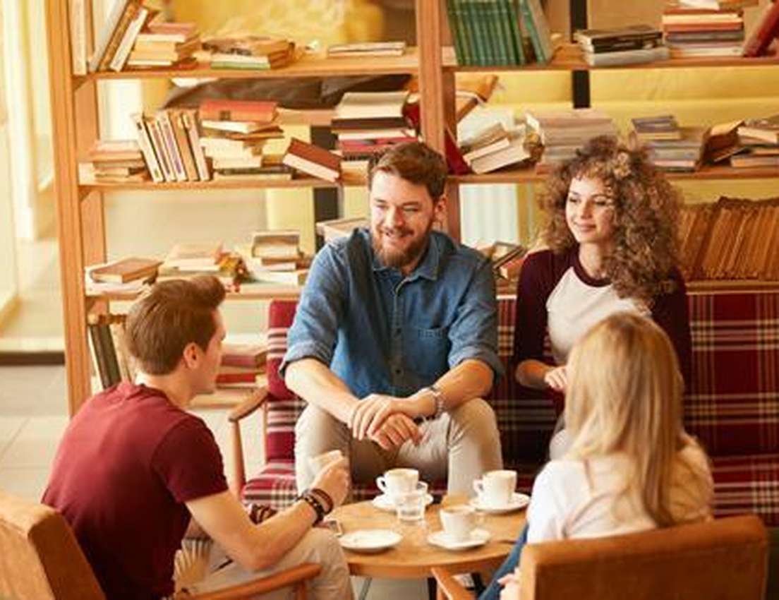 Group of four friends enjoying coffee and chatting in a cozy cafe with bookshelves in the background.