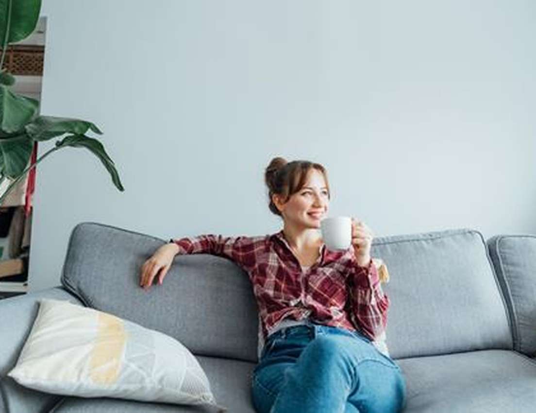 Woman in a plaid shirt sitting on a gray sofa, holding a mug, smiling, with a large plant and decorative pillows nearby.