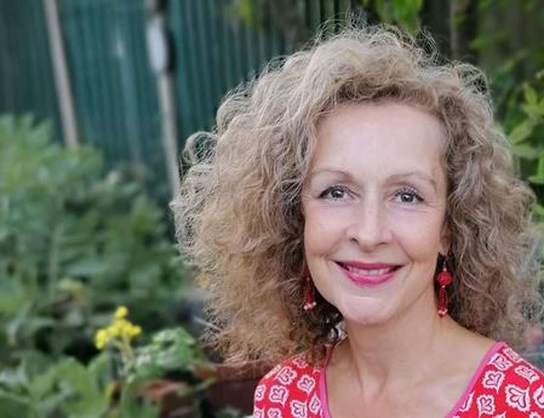 Smiling woman with curly hair and red earrings in a garden setting, wearing a red patterned blouse.