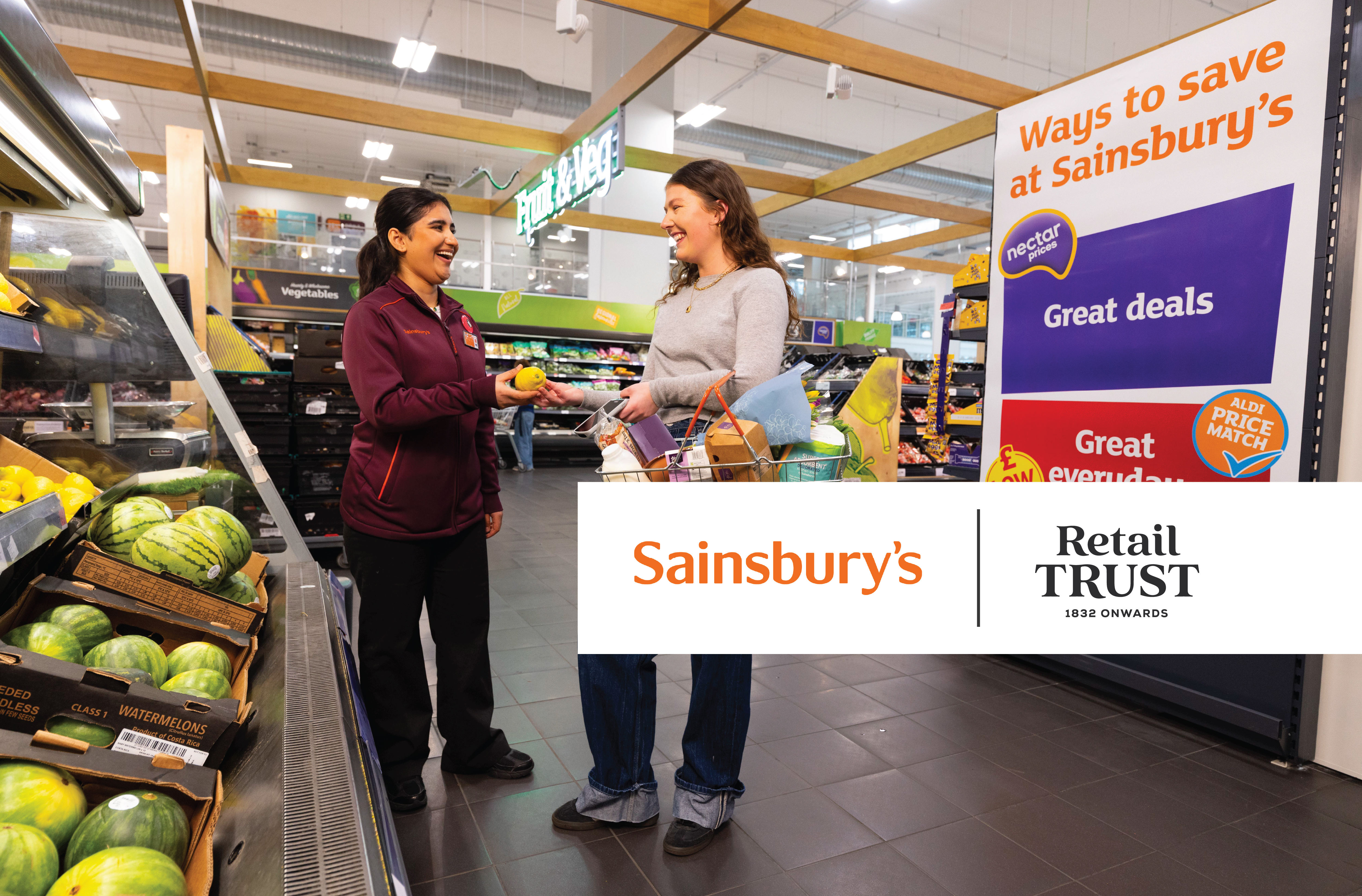 A Sainsbury's employee in a maroon top hands a yellow fruit to a smiling customer holding a shopping basket in the fruit and veg aisle.