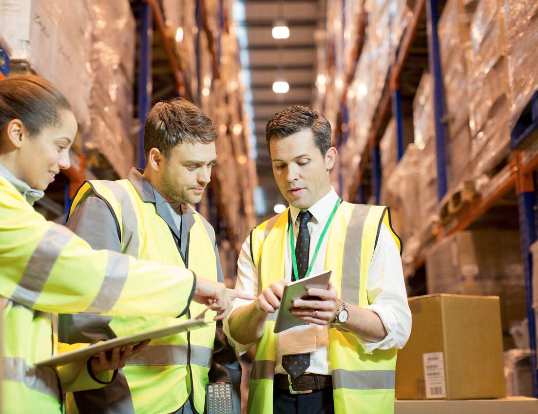 Three warehouse staff in high-vis vests review documents and a tablet amidst rows of stacked inventory.