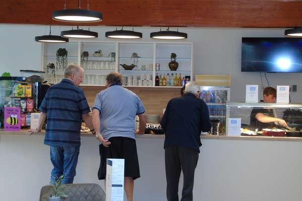 Three men stand at a modern cafe counter, with a woman serving food behind glass. Various drinks, snacks, and a menu are visi