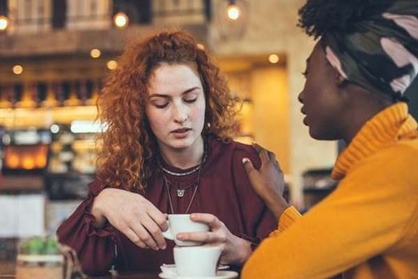 A woman with red curly hair looks down sadly, holding a coffee cup, as another woman comforts her with a hand on her shoulder in a warm cafe.