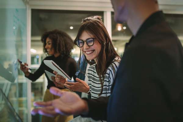 A group of diverse young professionals collaborating in an office. A smiling woman in glasses is central, while another writes on a glass board.