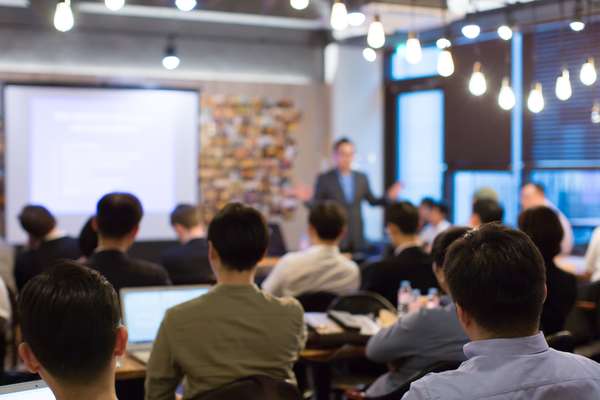 Blurred view of an audience from behind, facing a speaker and a projector screen in a brightly lit, modern conference room.