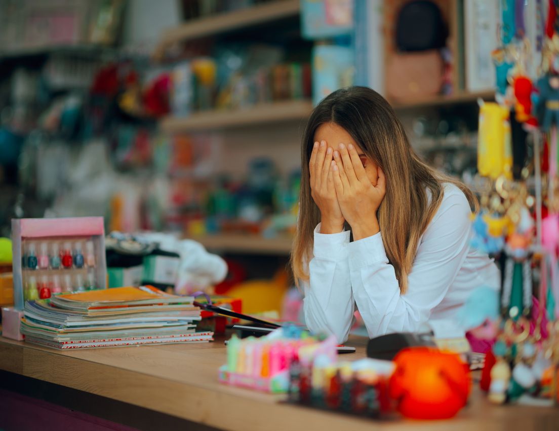 A woman with long hair covers her face with her hands, looking distressed or tired, at a colorful shop counter with books and